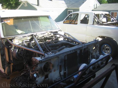 1990 Ford Bronco - stripped race truck with hood removed showing engine and frame, parked beside a white prerunner truck in a yard.
