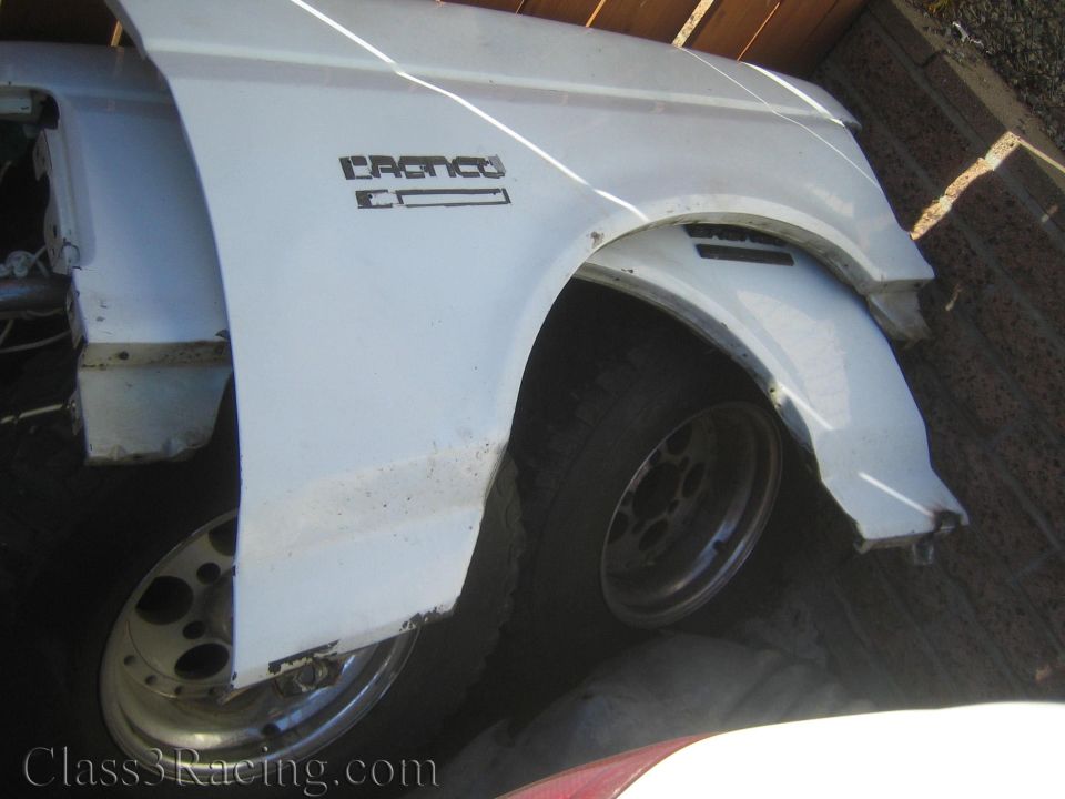 White Ford Bronco front fender lying over a wheel against a brick wall