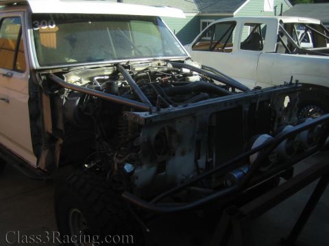 1990 Ford Bronco - exposed front end of a stripped race truck showing engine and frame rails, with a white truck parked behind.