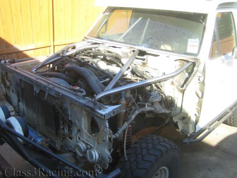 1990 Ford Bronco - stripped white truck cab with exposed engine, frame, and roll cage tubing parked against a wooden fence.