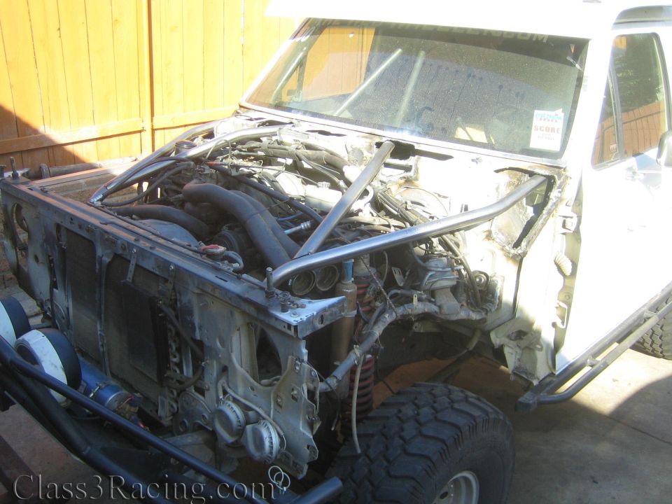 Stripped white truck cab with exposed engine and roll cage against wood fence