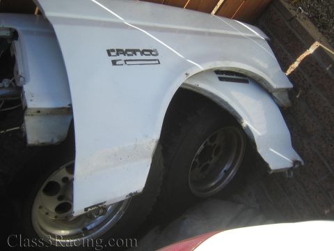 1990 Ford Bronco - white Ford Bronco front fender with embossed lettering rests on the ground beside off-road wheels and a brick wall.
