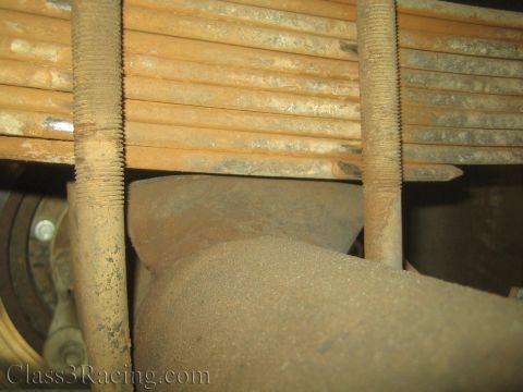 1990 Ford Bronco - rusted multi-leaf spring sits above a worn exhaust pipe in a vehicle undercarriage close-up.