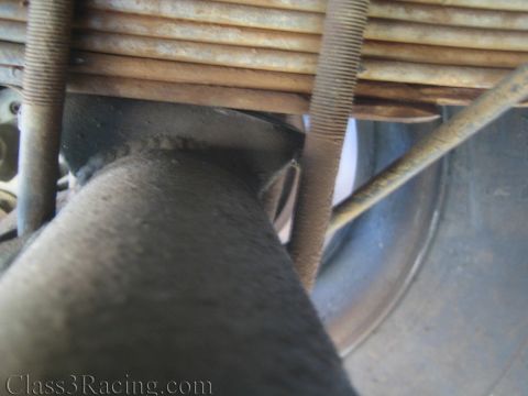 1990 Ford Bronco - dark exhaust pipe curves past a rusted leaf spring with U-bolts, near a chrome wheel rim underneath a vehicle.