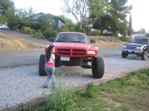 1990 Ford Bronco - woman crouches near the front of a red lifted Dodge Dakota pickup with oversized tires on a gravel lot.