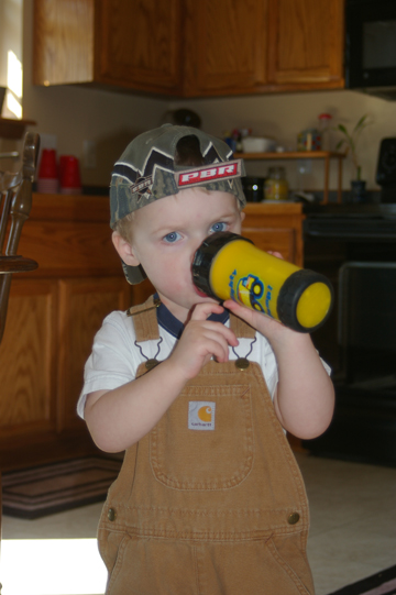 1998 Jeep TJ - A young child wearing overalls and a PBR hat is seen drinking from a yellow cup in a kitchen setting.