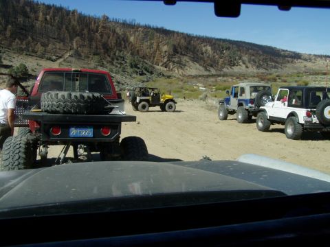 1967 Ford Bronco - A group of off-road vehicles are parked in a dusty, mountainous area, with a red pickup truck in the foreground.