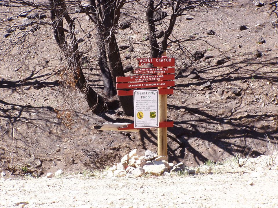 Red trail sign with text about Jacket Canyon and a pledge, set against a dry, rocky landscape with burnt trees