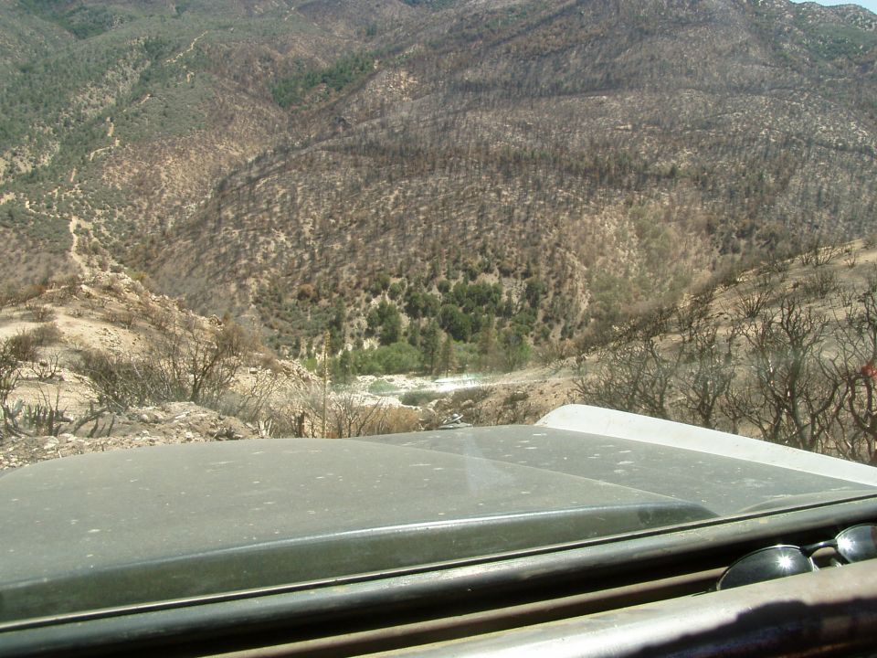 Green vehicle hood with a roll bar and sunglasses, overlooking a dry, mountainous landscape with sparse vegetation