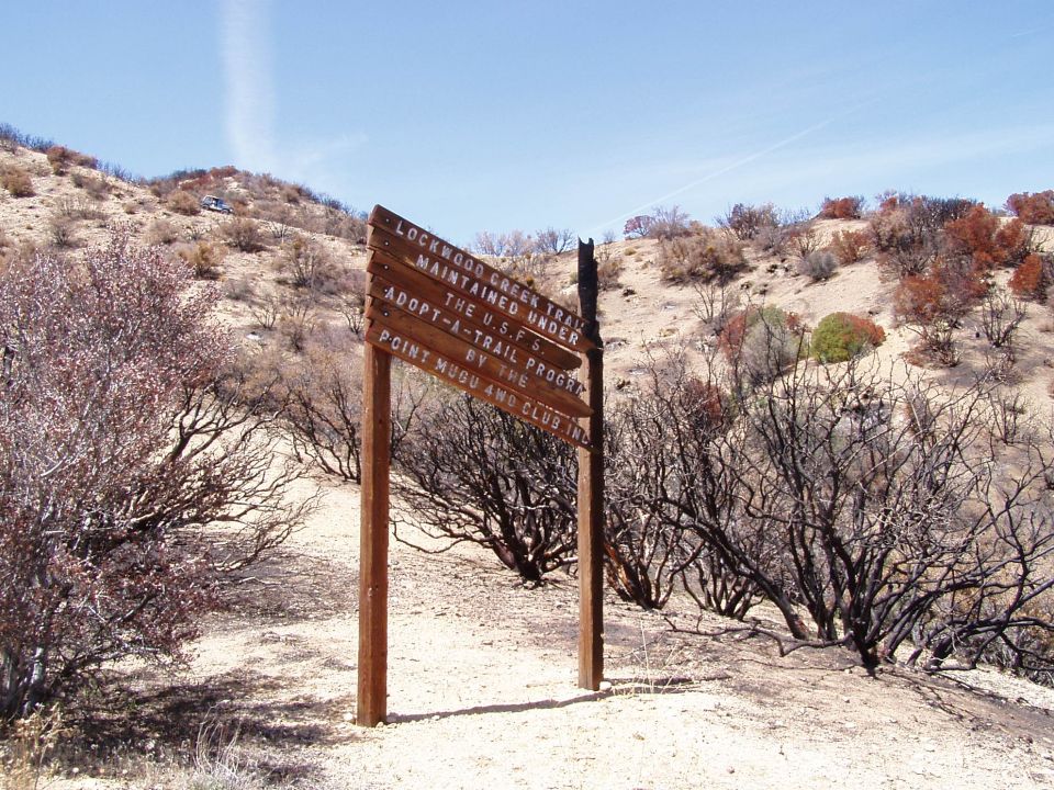 Wooden trail signs with text about Lockwood Creek Trail and Adopt-A-Trail Program, set against a dry, hilly landscape with sparse vegetation