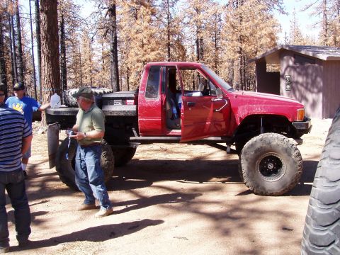 1967 Ford Bronco - A modified red pickup truck with large off-road tires is parked in a wooded area with several people nearby.
