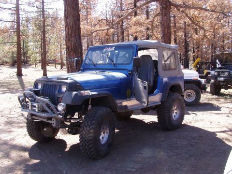 1967 Ford Bronco - A blue Jeep Wrangler is parked in a wooded area with other vehicles visible in the background.