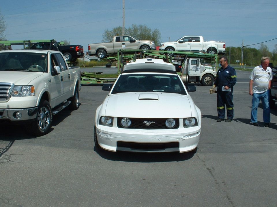 White Ford Mustang with tape on the front bumper, parked next to a white Ford F-150 pickup truck. Two men stand nearby