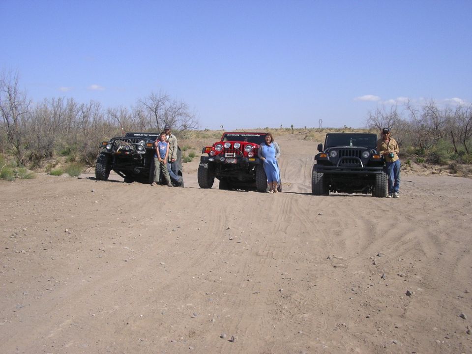 Three lifted off-road vehicles parked on a sandy desert trail with people standing beside them