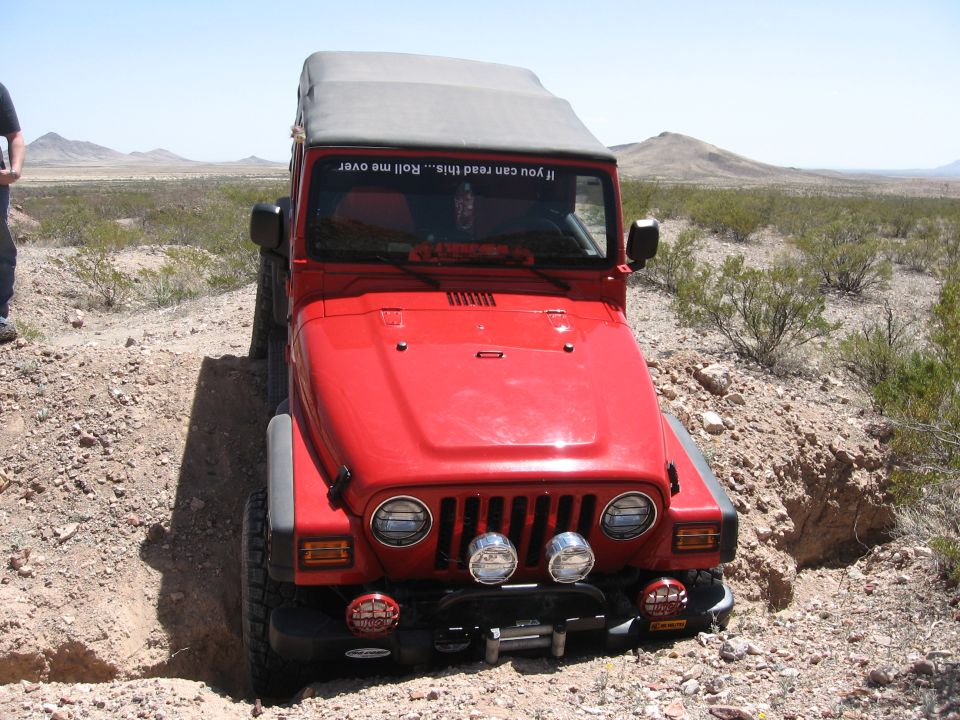 Red Jeep Wrangler on dirt trail, front view. Sticker on windshield says 'If you can read this... Roll me over'