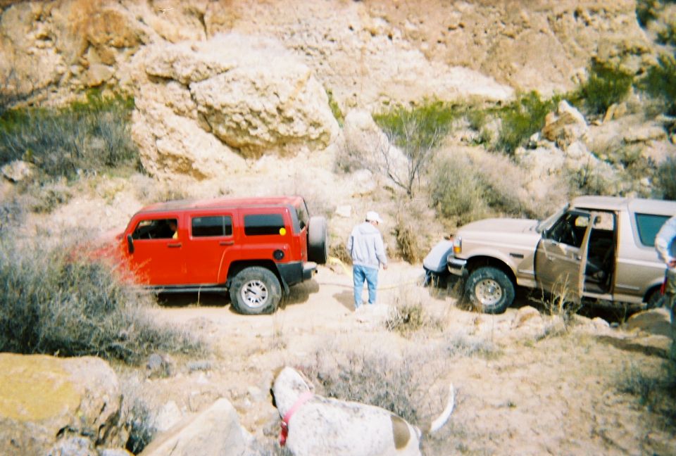 Red 4-door SUV towing a tan SUV with a strap on a rocky desert trail; man standing between them, dog in foreground