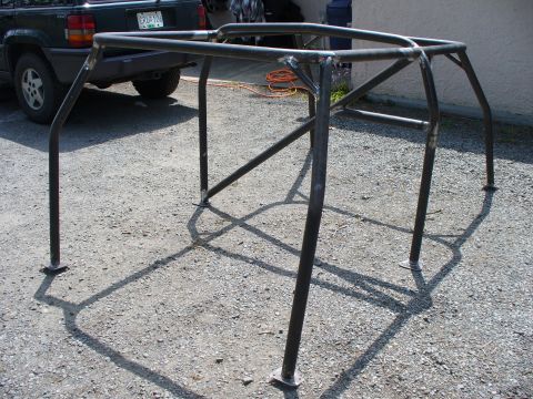 1978 Ford Bronco - A custom-built roll cage frame sits on a gravel surface, with a dark SUV visible behind it.