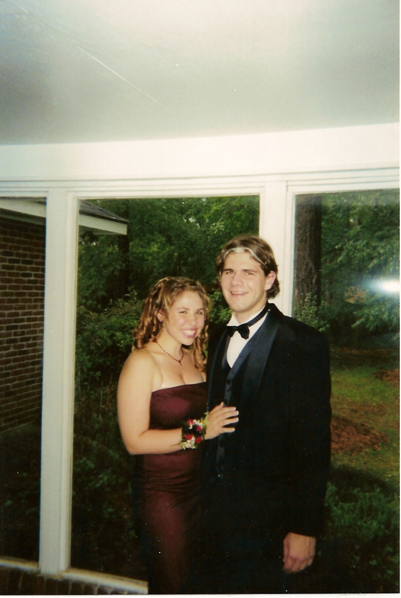 Couple in formal attire posing for a photo indoors with a brick wall and trees visible through a window