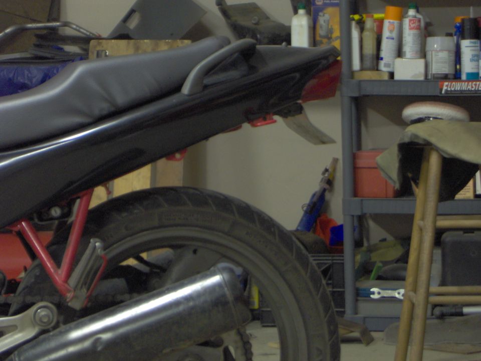 Motorcycle rear seat, tail light, and exhaust pipe visible. Shelving with supplies in background