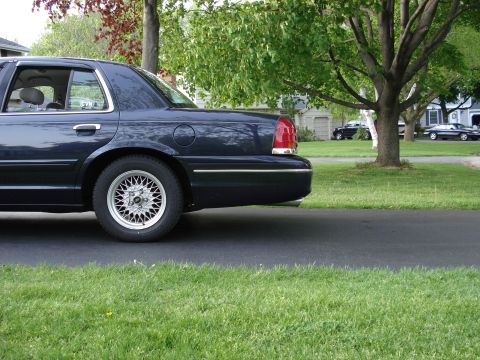 2000 Ford Crown Victoria - Low side view of the rear half of a dark blue full-size Ford sedan sitting on a driveway apron, framed against a green suburban lawn and sh…
