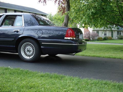 2000 Ford Crown Victoria - Low ground-level rear three-quarter view of a dark navy blue full-size four-door sedan parked on a residential driveway, shot from behind t…