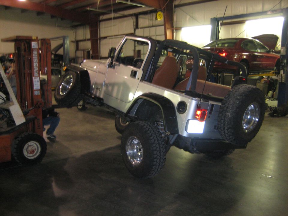 Silver 2002 Jeep Wrangler lifted by a forklift in a garage; another car is on a lift in the background