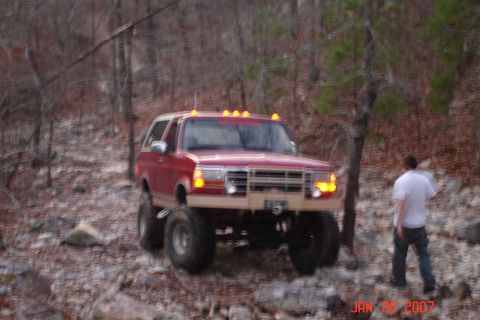 1995 Ford Bronco - A lifted red Ford Bronco navigates a rocky, wooded terrain with a person standing nearby.