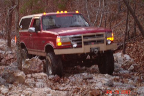 1995 Ford Bronco - This red 1995 Ford Bronco navigates a challenging rocky terrain, equipped with a tan aftermarket bumper and amber roof lights.