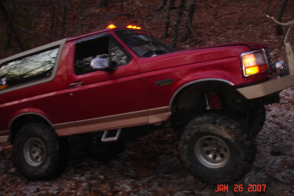 Side view of lifted red and tan Ford Bronco climbing rocks, red shocks and rock sliders visible