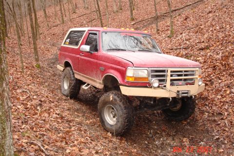 1995 Ford Bronco - This red 1995 Ford Bronco is equipped for off-road adventures, navigating a muddy trail through a forest.