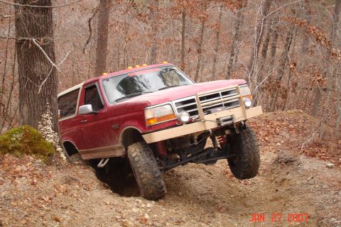 1995 Ford Bronco - A lifted red Ford Bronco with a tan brush guard, winch and tan rocker guards flexes hard over a dirt embankment in bare winter woods, front…