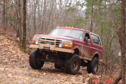 1995 Ford Bronco - Three-quarter front view of a lifted red Ford Bronco with a tan removable hardtop, flexing its front suspension on a rocky leaf-covered hil…