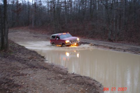 1995 Ford Bronco - A red Ford Bronco with a white top and amber cab roof lights blasts through a long muddy brown water hole on a dirt trail at dusk, throwing…