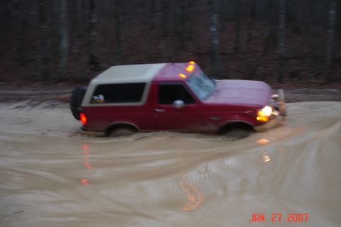 1995 Ford Bronco - A maroon Ford Bronco with a white fiberglass top and cab-roof marker lights wades through deep muddy brown water at dusk, headlights on and…