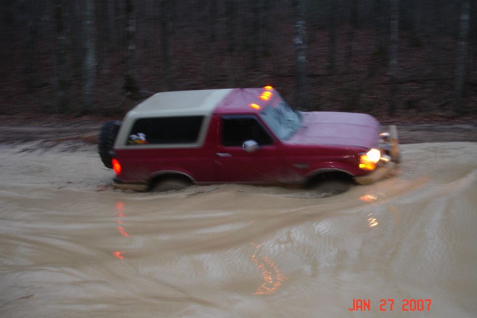Maroon Ford Bronco with white top wading through deep muddy water at dusk