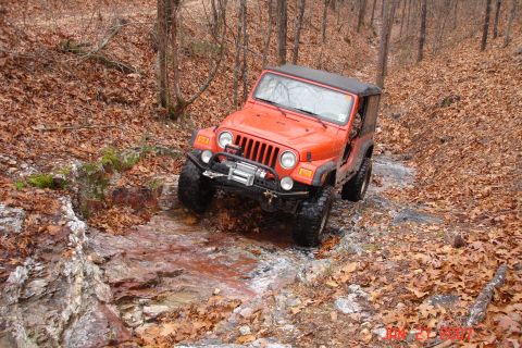 1995 Ford Bronco - Orange Jeep Wrangler navigates a stream in a wooded area during autumn.