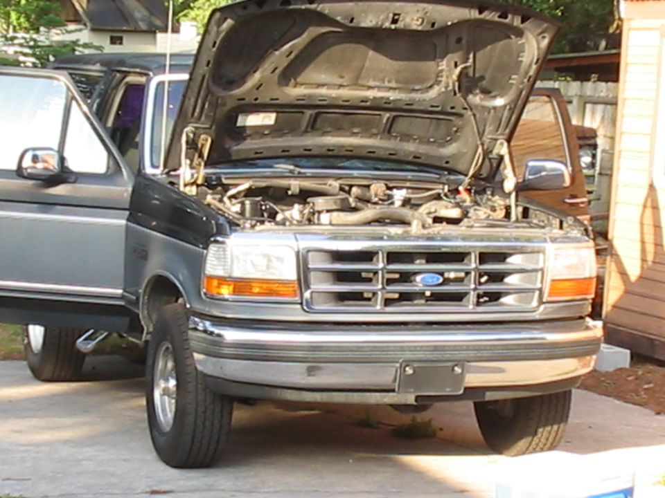Front view of a dark gray and silver Ford F-Series truck with hood open, showing engine. Another vehicle is partially visible behind it