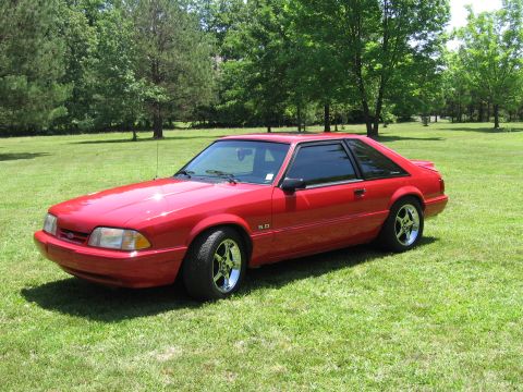 1967 Ford Mustang - A red Ford Mustang is parked on a grassy lawn with trees in the background.