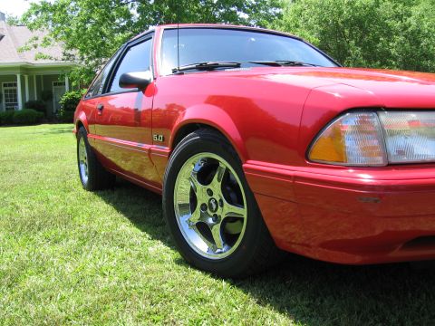 1967 Ford Mustang - Red coupe with 5.0 badge and chrome wheels parked on grass in front of a house.