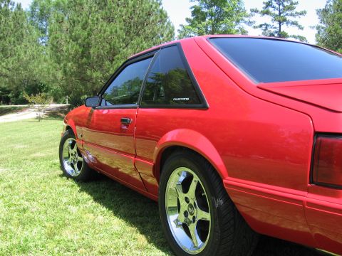 1967 Ford Mustang - A red coupe with tinted windows is parked on a grassy area with trees in the background.