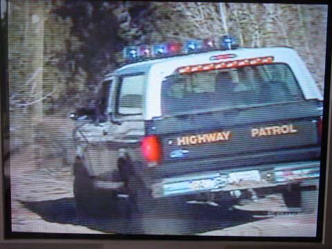 1994 Ford Bronco - Photograph of a TV or monitor screen showing the rear of a black-and-white Highway Patrol SUV with a roof-mounted light bar, amber rear dec…