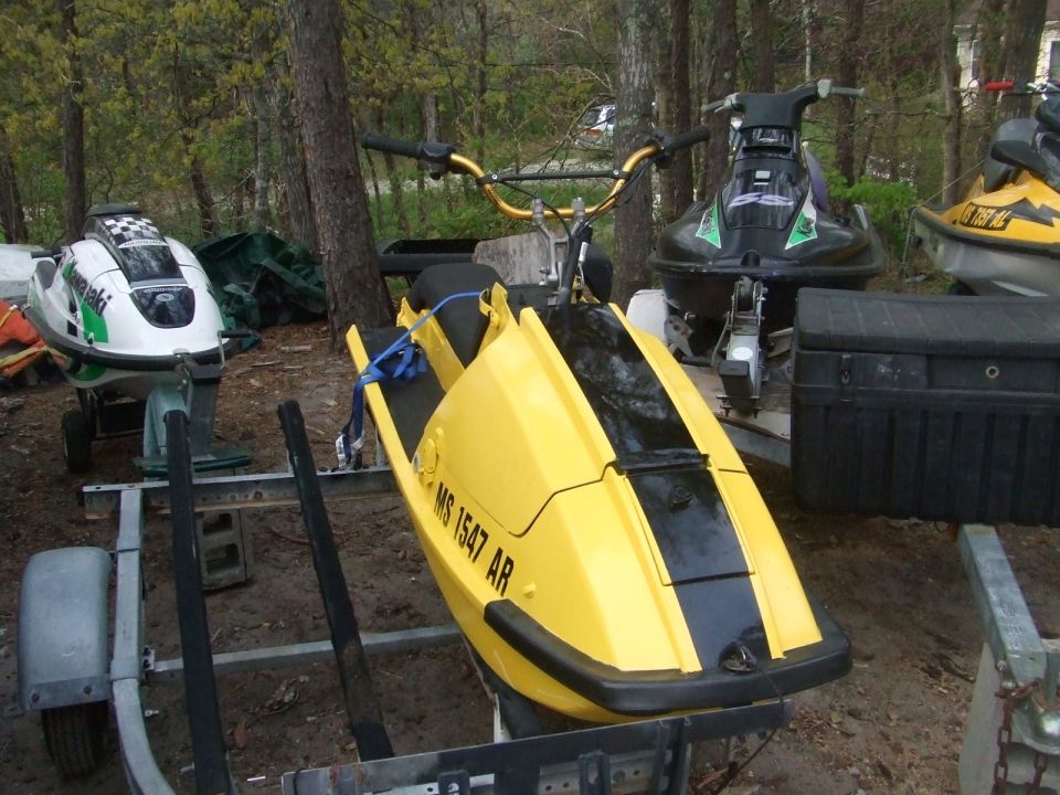 Three jet skis on trailers in a wooded area. One is yellow and black with 'MS 1547 AR' on the side