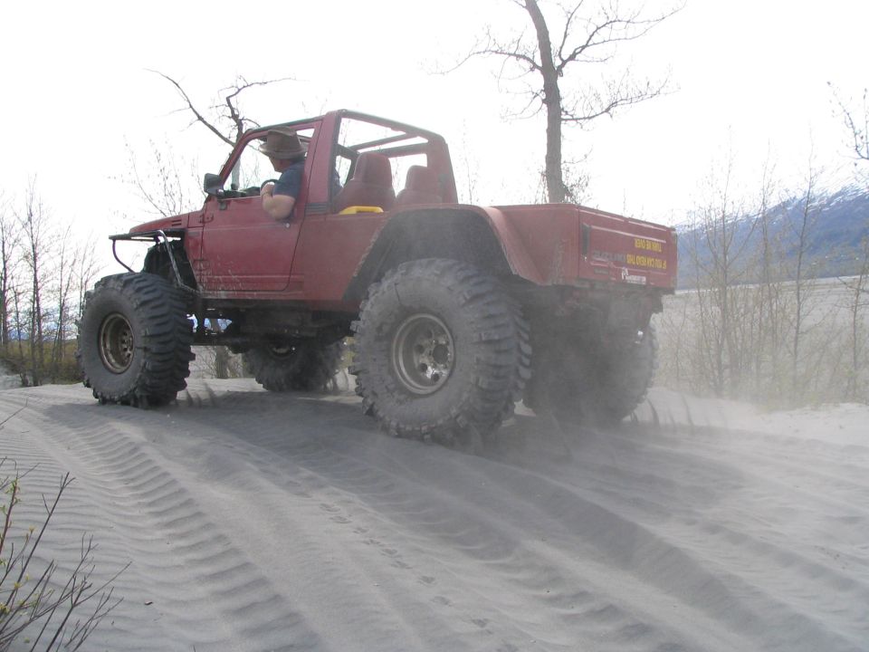Red topless 4x4 on huge mud tires driving up a sand dune, driver in straw hat