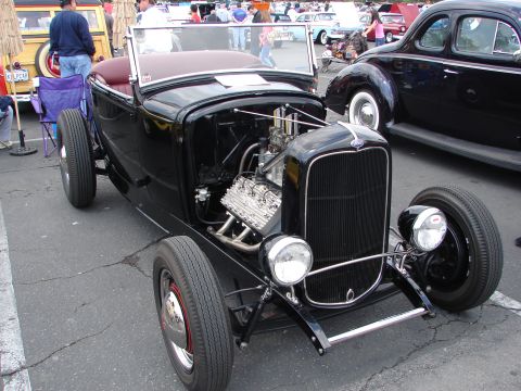 1992 Ford Taurus - A black hot rod roadster with a prominent V8 engine is displayed at a car show, with other vehicles visible in the background.