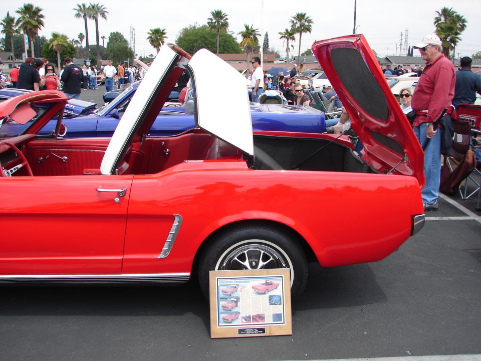 Red 1965 Ford Mustang convertible with white top up, hood and trunk open. Blue car behind. Display board in front of wheel