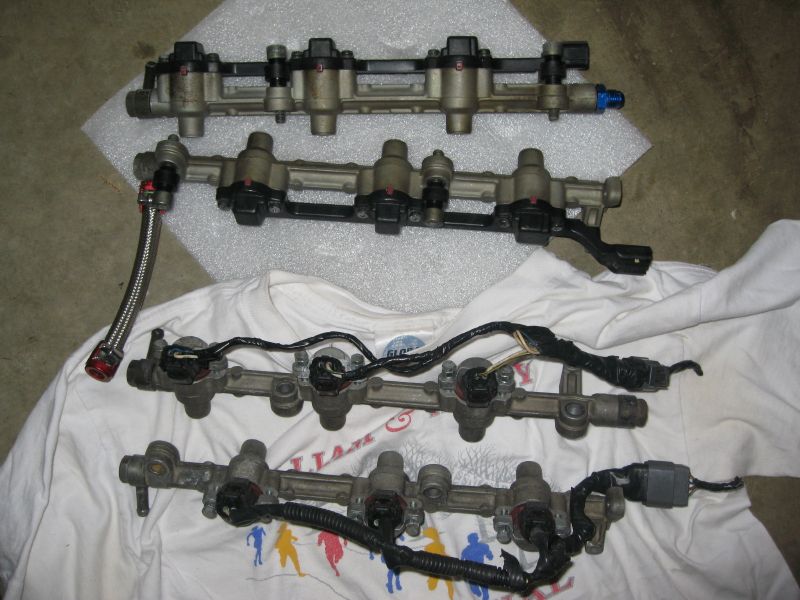 Four fuel injector rails with injectors and fittings on a white shirt and concrete floor