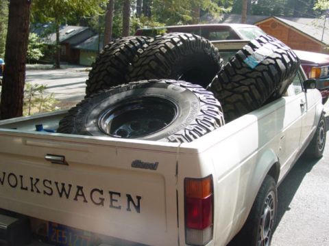 1986 Ford Bronco - A white Volkswagen pickup truck carries large off-road tires in its bed, with other vehicles and houses in the background.