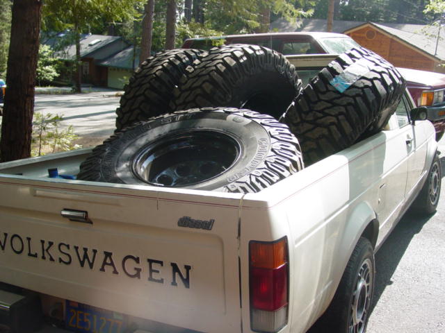 Volkswagen pickup truck bed loaded with three large off-road tires. The truck is white