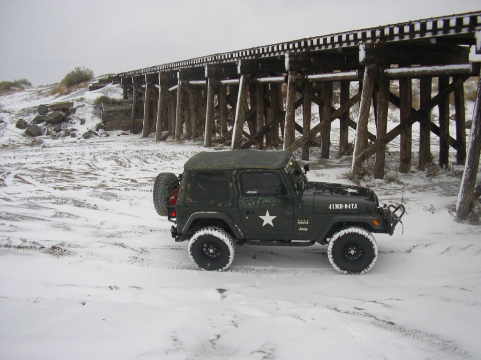 Green Jeep Wrangler with a white star on the side parked in the snow under a wooden trestle bridge. License plate reads 41MB-04TJ