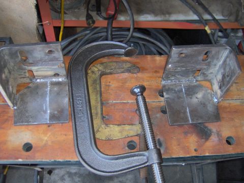 1993 Ford Bronco - Metal brackets are held in place with clamps on a wooden workbench, with welding visible on the brackets.